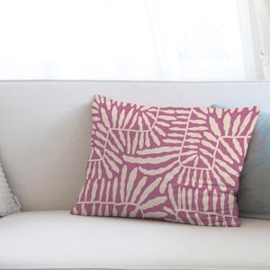 May include: A decorative pillow with a pink background and a cream-colored, abstract leaf pattern. The pillow is rectangular and rests on a white sofa. The pillow's design adds a touch of modern style to home decor.
