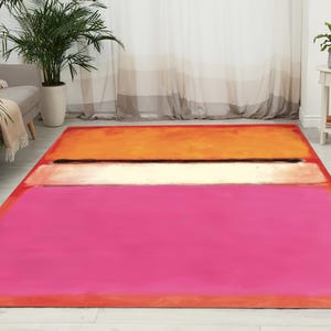 Puede incluir: Una alfombra rectangular con un diseño abstracto moderno. La alfombra presenta tres bandas horizontales de color: un rosa vibrante en la parte inferior, una banda de color crema en el medio y una banda naranja en la parte superior. La alfombra está colocada sobre un suelo de madera clara.