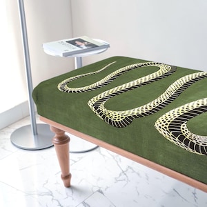 May include: A rectangular bench featuring a dark olive green fabric upholstery with a large, winding snake graphic in yellow and black. The bench is supported by four turned wooden legs with a light brown finish, showcasing a classic furniture design.