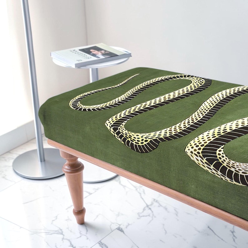 May include: A rectangular bench featuring a dark olive green fabric upholstery with a large, winding snake graphic in yellow and black. The bench is supported by four turned wooden legs with a light brown finish, showcasing a classic furniture design.