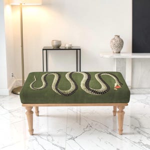 May include: A rectangular ottoman featuring a dark olive green fabric top with a large, winding snake graphic in black and cream with a red tongue. The piece is supported by four light-toned wooden legs with a turned, spindle-style design.
