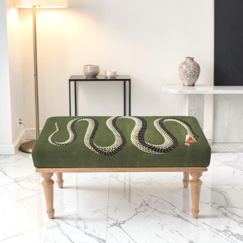 May include: A rectangular ottoman featuring a dark olive green fabric top with a large, winding snake graphic in black and cream with a red tongue. The piece is supported by four light-toned wooden legs with a turned, spindle-style design.