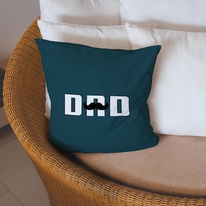 May include: Teal throw pillow with the word "DAD" in white block letters and a black mustache graphic. The pillow is on a wicker chair with white pillows in the background.