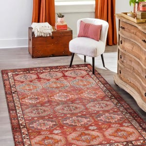 Peut inclure: Un tapis rectangulaire rouge avec un motif géométrique complexe. Le tapis présente un motif central de médaillons répétitifs dans des tons de rouge, d'orange et de gris, encadré par une bordure détaillée. Le tapis est posé sur un sol en bois gris clair.