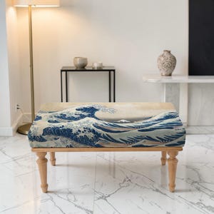 May include: A wooden ottoman with a rectangular top featuring a print of Hokusai's "The Great Wave off Kanagawa." The artwork depicts a large ocean wave in shades of blue and white, with a beige background. The ottoman has four turned wooden legs.