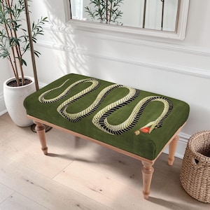 Pode incluir: Banqueta retangular com tampo estofado verde com uma ilustração detalhada de uma cobra. O desenho da cobra tem segmentos pretos e dourados com uma língua vermelha. A banqueta tem pernas de madeira de cor clara.