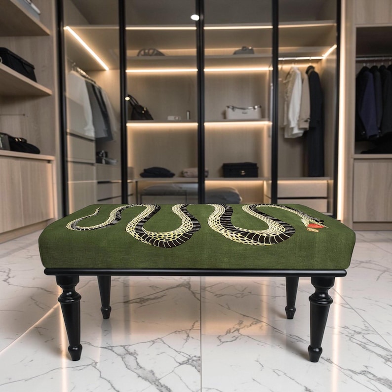 May include: A rectangular ottoman featuring a green fabric top with a black and yellow snake illustration. The piece is supported by four black wooden legs with a turned design, providing a stable base for this furniture item in a room.