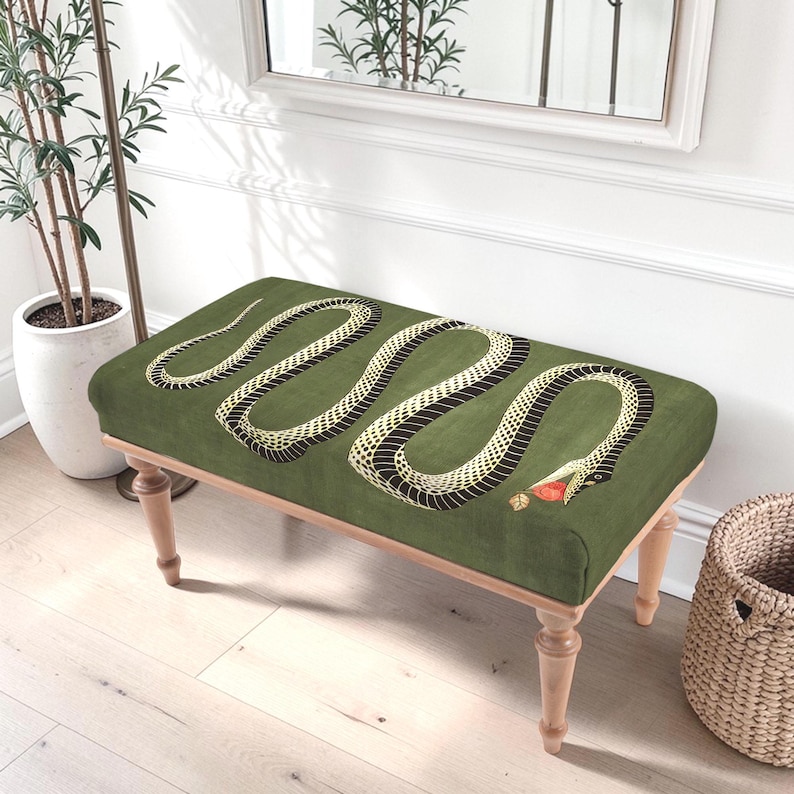 May include: A rectangular bench with a moss green upholstered top featuring a large, winding black and cream-colored snake graphic. The snake has an open mouth holding a small red apple. The bench is supported by four light-toned wooden legs.