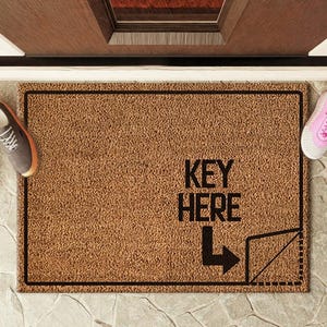 Può includere: Un tappetino per porta in fibra di cocco marrone con testo nero che dice "KEY HERE" e una freccia che punta a sinistra. Il tappetino è su un pavimento di pietra con scarpe da ginnastica rosa su entrambi i lati.