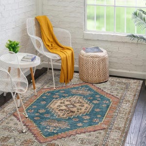 May include: A patterned area rug featuring a central teal and beige geometric design, with a rust-coloured trim and a neutral patterned edge. The rug is in a room with white chairs, a small table, and a woven ottoman.