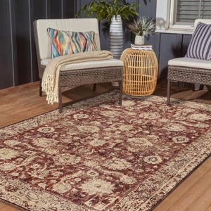 May include: A rectangular area rug with a distressed, floral pattern in burgundy and beige tones. The rug is placed on a wooden deck, with outdoor furniture and decorative pillows visible in the background. The rug's design features a detailed border.