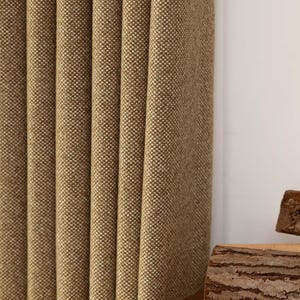 May include: A close-up of a textured, tan curtain with vertical folds. A wooden table holds a book with the text "ALVAR AALTO" and two pieces of wood. The background is a white wall.