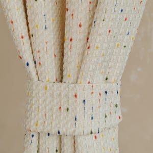 Può includere: Tenda color crema con tessuto strutturato, ornata da piccoli punti colorati rossi, gialli, blu e verdi. La tenda è raccolta e tenuta da un fermatenda in tessuto abbinato, creando un drappeggio decorativo.