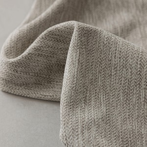 May include: Close-up of a folded, textured fabric in shades of gray and beige. The fabric has a herringbone weave pattern, with the threads creating a subtle, diagonal design. The material appears soft and pliable.