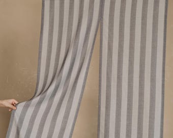 Puede incluir: Panel de cortina a rayas grises y blancas colgado de una barra negra. La tela tiene un patrón de rayas verticales. La cortina está hecha de un material suave y tejido. La cortina se muestra sobre un fondo neutro.