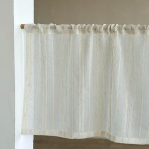 Può includere: Un pannello di tenda a righe color crema è appeso a un'asta di legno. La tenda ha strisce verticali ed è realizzata in un tessuto leggero e strutturato. La tenda copre parzialmente una finestra o una porta.
