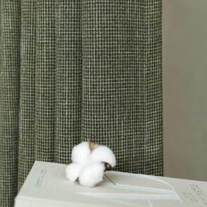 May include: A dark olive green curtain with a fine white grid pattern hangs vertically. In the foreground, a white cotton boll rests on a light beige book titled AD at 100 A CENTURY OF STYLE, which is stacked on a brown surface.