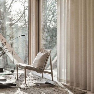 May include: A bright room featuring a wooden chair, a floor lamp, and neutral curtains. A window provides a view of trees. A book and a cushion are on the floor. The room has a minimalist, natural aesthetic.