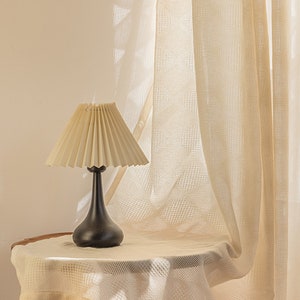 Puede incluir: Una l&aacute;mpara de mesa negra con una pantalla plisada de color crema se encuentra sobre una mesa blanca con una cortina blanca transparente en el fondo.