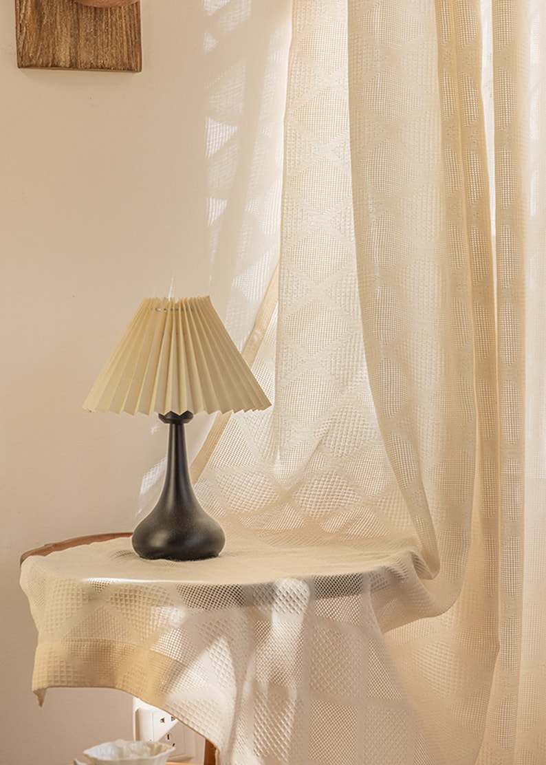 Puede incluir: Una l&aacute;mpara de mesa negra con una pantalla plisada de color crema se encuentra sobre una mesa blanca con una cortina blanca transparente en el fondo.