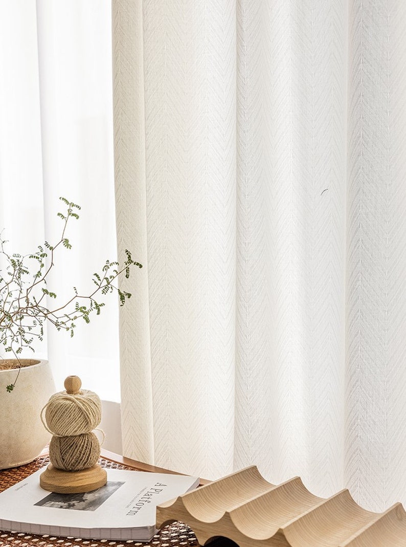 May include: White sheer curtains with a subtle herringbone pattern hang in a window. A small potted plant and a wooden spool of twine are on a table in front of the curtains.