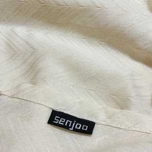 May include: Close-up of a white fabric with a textured pattern. A black label with the word "senjoo" is sewn onto the fabric.
