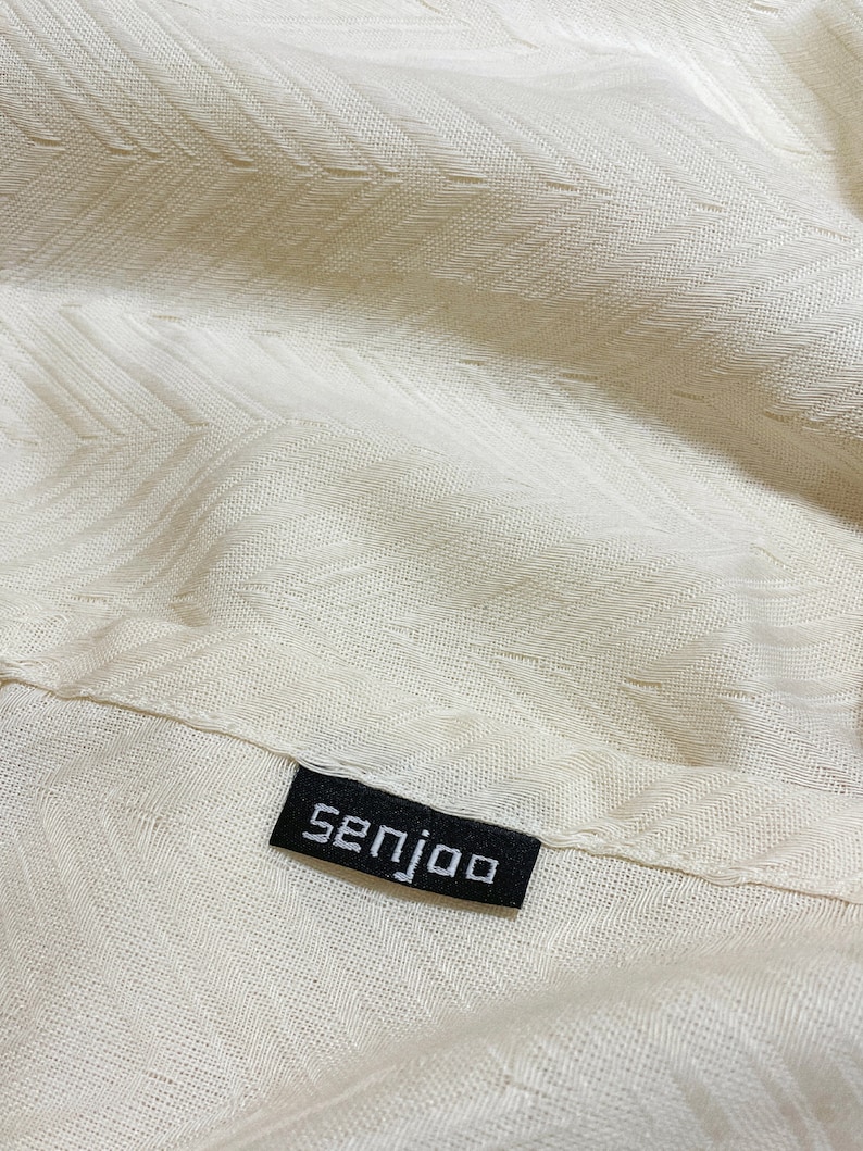 May include: Close-up of a white fabric with a textured pattern. A black label with the word "senjoo" is sewn onto the fabric.