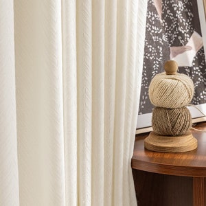 May include: White sheer curtains with a subtle textured pattern hang in front of a window. A wooden spool with three balls of twine sits on a wooden table.