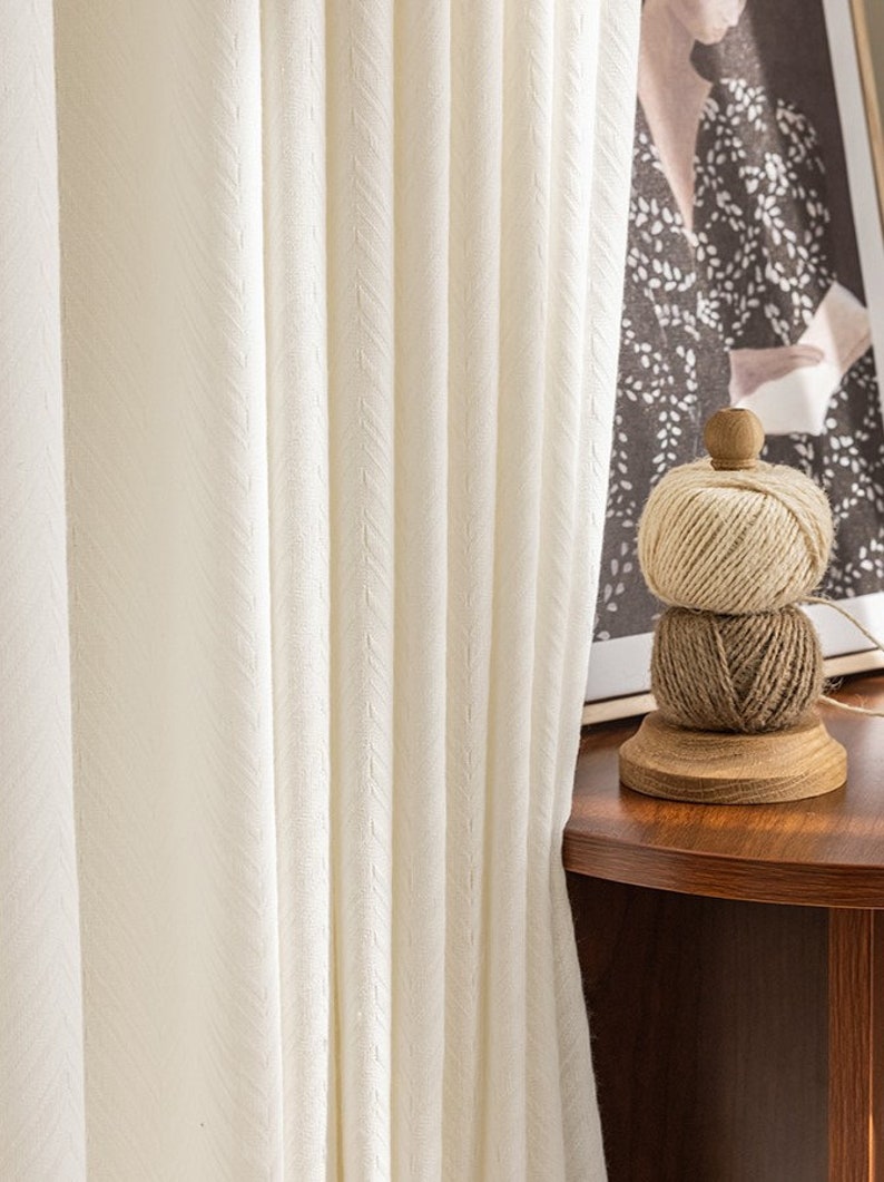 May include: White sheer curtains with a subtle textured pattern hang in front of a window. A wooden spool with three balls of twine sits on a wooden table.