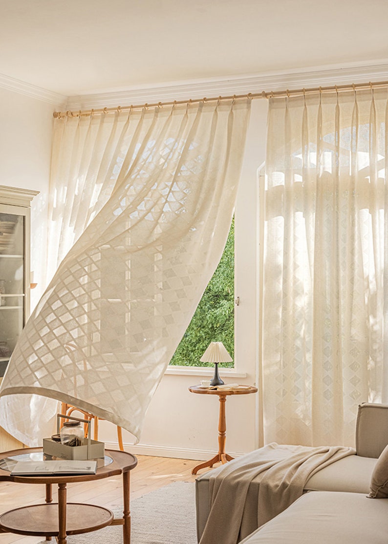 Puede incluir: Cortinas blancas y transparentes con un patr&oacute;n geom&eacute;trico cuelgan en una sala de estar con una mesa de madera y una l&aacute;mpara. Las cortinas se agitan con el viento.