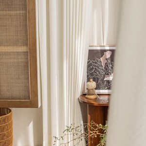 May include: White curtains with a wooden side table and a framed print of a woman in a black and white dress. The table has a spool of twine on it.