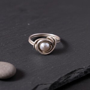 Könnte beinhalten: Ein Ring aus silberfarbenem Draht mit einer einzelnen runden weißen Perle in der Mitte. Der Metalldraht ist um die Perle gewickelt und bildet ein geschichtetes, kreisförmiges Design, das sowohl die Ringschiene als auch die Fassung bildet.