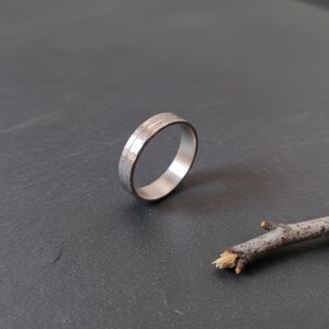 Könnte beinhalten: Ein silberfarbener Metallring mit einer strukturierten, rindenartigen Oberfläche auf der Außenseite. Das Band hat eine glatte, polierte Innenseite und eine gleichmäßige Breite, die ein detailliertes, raues Muster aufweist, das an Holzmaserung erinnert.