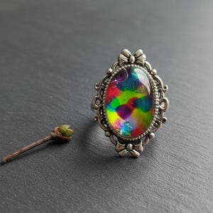 May include: A silver-toned ring featuring an oval-shaped centerpiece with a vibrant, multi-colored tie-dye pattern of red, green, blue, and yellow, surrounded by an ornate, filigree-style metal frame with small bow-like details at the top and bottom.