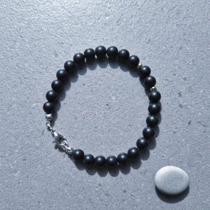 Könnte beinhalten: Ein schwarzes Perlenarmband mit silbernem Verschluss und Akzenten. Das Armband besteht aus kleinen, runden, mattschwarzen Perlen. Ein kleiner, glatter, grauer Stein liegt neben dem Armband.