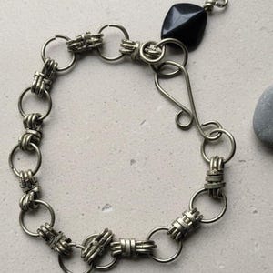 Könnte beinhalten: Ein silberfarbenes Armband mit einer Reihe von miteinander verbundenen Ringen und dekorativen Gliedern. Das Armband hat eine schwarze, quadratische Perle an einem Ende und einen Hakenverschluss. Der Schmuck wird auf einer neutralen Oberfläche präsentiert.