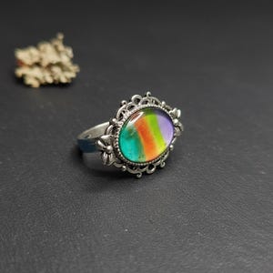 May include: A silver-toned ring featuring an oval-shaped cabochon with a gradient of purple, green, and orange colors. The metal setting has an intricate, decorative filigree border with small floral-like details on the sides of the band.