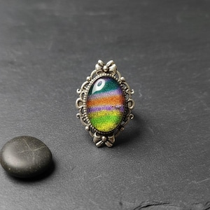 May include: A silver-toned ring featuring an oval-shaped centerpiece with a multi-colored, glittery gradient pattern in shades of green, purple, and orange. The metal setting has an intricate, decorative filigree border with small bead accents.