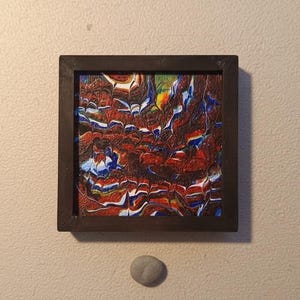 Könnte beinhalten: Quadratisches abstraktes Gemälde in einem dunkelbraunen Rahmen. Das Kunstwerk zeigt eine Mischung aus roten, blauen, weißen und gelben Farben. Unter dem Gemälde befindet sich ein kleiner, grauer, ovaler Stein an einer beigen Wand.