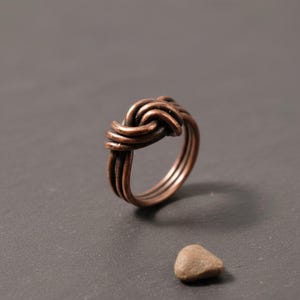 Könnte beinhalten: Ein Kupferring mit einem mehrsträngigen Drahtdesign, das sich zu einem zentralen Knoten windet. Das Metall hat ein dunkles, oxidiertes Finish mit sichtbarer Textur entlang der gewickelten Drähte, was dem Ring ein geschichtetes Aussehen verleiht.