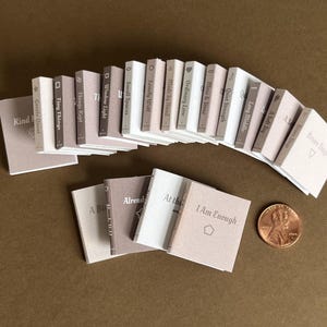 Peut inclure: Une collection de petits livres miniatures aux tons beige et marron, disposés sur une surface marron. Les titres incluent "I Am Enough" et "Tiny Things". Une pièce de monnaie est incluse pour l'échelle.