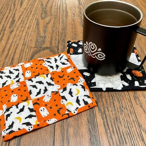 May include: Two Halloween-themed fabric coasters. One coaster is orange and white with a pattern of ghosts, bats, and crescent moons. The other coaster is black and white with a pattern of ghosts, bats, and spiderwebs. A black mug with a white design is sitting on top of the black and white coaster.