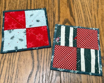 Op de afbeelding: Twee handgemaakte gequilte onderzetters met een kerstthema. De ene onderzetter heeft een rood en wit sneeuwvlokkenpatroon en een wit en grijs dennennaaldenpatroon. De andere onderzetter heeft een rood en wit ruitjespatroon en een wit en grijs berkenboompatroon.