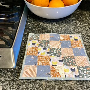May include: A patchwork quilted hot pad with a blue border. The patchwork design features various floral and geometric patterns in shades of brown, blue, and yellow. The hot pad is made of cotton fabric and is perfect for protecting surfaces from heat.