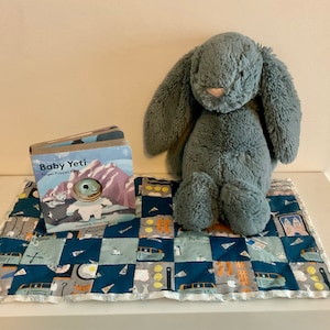 May include: A blue and grey baby blanket with a patchwork design. The blanket features a variety of patterns, including cars, stars, and geometric shapes. A grey plush bunny and a book titled "Baby Yeti Finger Puppet Book" are on top of the blanket.