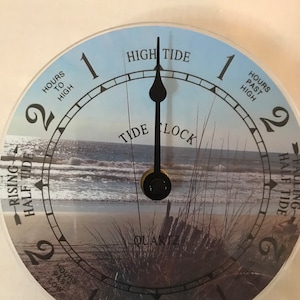 May include: A white and blue tide clock with a black hand pointing to the high tide mark. The clock face features a beach scene with waves, sand, and seagrass. The text "TIDE LOCK" is printed on the clock face.