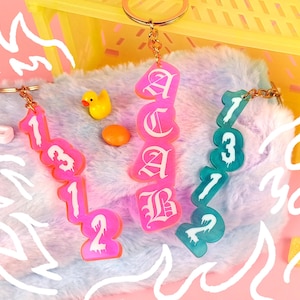 Può includere: Tre portachiavi con design diversi. Un portachiavi è rosa con i numeri 1312, un altro è verde acqua con i numeri 1312 e l'ultimo è rosa con le lettere CAB in carattere gotico.