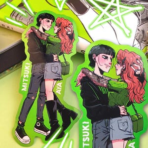 Może przedstawiać: Dwa naklejki przedstawiające parę w zielono-czarnej kolorystyce. Naklejki znajdują się na zielonym tle z białym tekstem, który brzmi "Mitsuki" i "Aya".