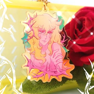 Puede incluir: Un amuleto de metal dorado con una ilustración de dibujos animados de una mujer con el pelo largo y rubio, que lleva una corona de espinas. El fondo es un degradado rosa y naranja.