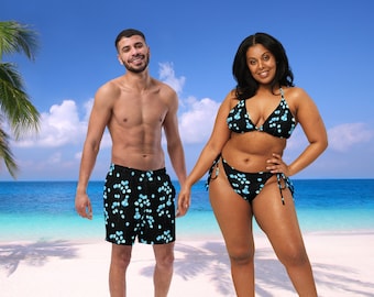 May include: Two people wearing matching black and blue swimwear with a floral pattern. The swimwear has a tropical vibe. The people are standing on a sandy beach with a blue ocean in the background.